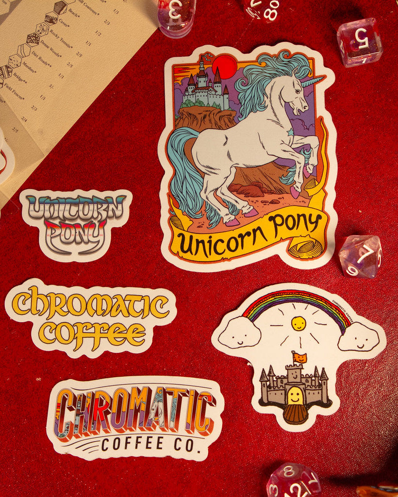 A collection of colorful stickers with unicorn and castle illustrations, branding for Chromatic Coffee, and other related designs laid out on a red surface.