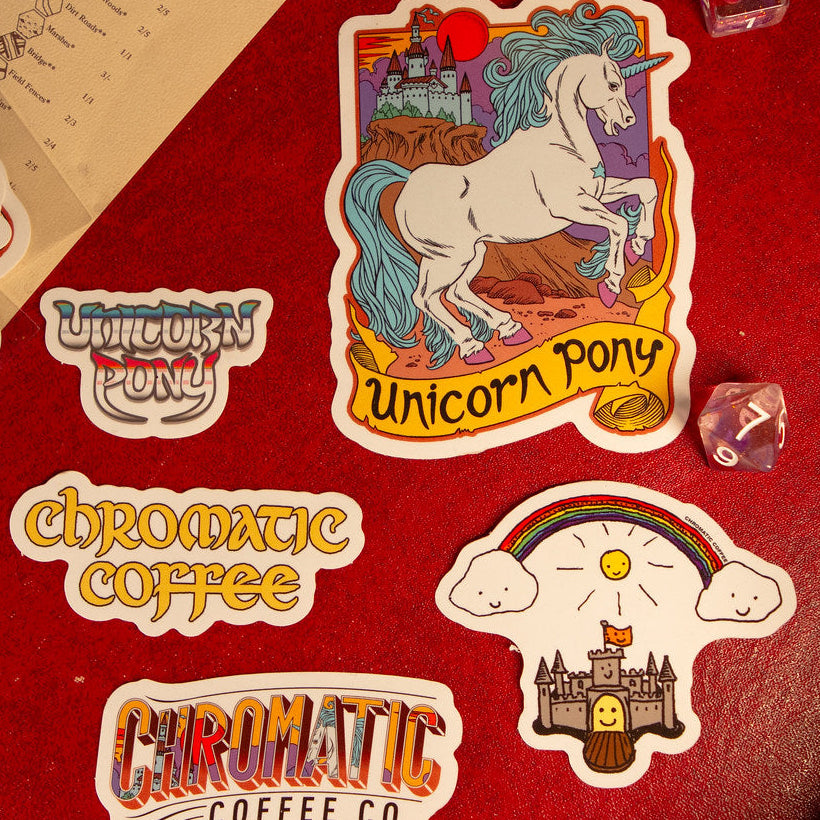 A collection of colorful stickers with unicorn and castle illustrations, branding for Chromatic Coffee, and other related designs laid out on a red surface.